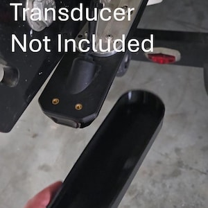 May include: A black transducer mount with a silver bracket is attached to a boat. The text "Transducer Not Included" is visible. A black cover is held in the foreground.