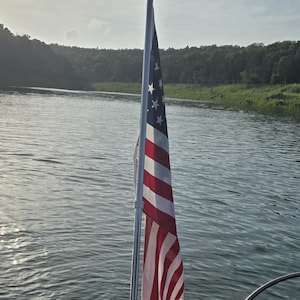 May include: A vertical metal flag pole holding a United States flag with red, white, and blue stripes and white stars on a blue field. The text "Flag pole, flag and boat not included." is visible at the top of the image against the sky.