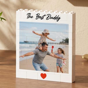 Puede incluir: Marco de fotos de bloques blancos con una foto familiar en la playa. El marco dice "The Best Daddy" en escritura negra, con un corazón rojo en la parte inferior. A la izquierda hay un vaso de agua con limón y vegetación.