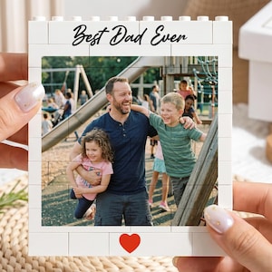 Puede incluir: Marco de fotos blanco en forma de bloque con una foto familiar en el interior. El marco tiene las palabras "Best Dad Ever" y un corazón rojo. La foto muestra a un hombre con dos niños en un parque infantil.