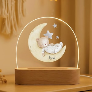 Puede incluir: Una luz de noche con un oso de peluche durmiendo en una luna creciente con estrellas. El diseño está impreso en un panel acrílico transparente, iluminado por una luz LED cálida. La base de madera tiene el nombre "James".