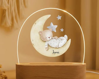 Puede incluir: Una luz de noche con un oso de peluche durmiendo en una luna creciente con estrellas. El diseño está impreso en un panel acrílico transparente, iluminado por una luz LED cálida. La base de madera tiene el nombre "James".