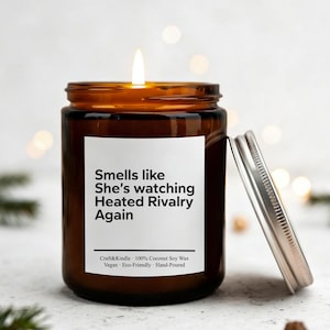 Peut inclure: Un pot de bougie en verre brun allumé avec un couvercle argenté, avec le texte "Smells like She's watching Heated Rivalry Again". La bougie est entourée d'aiguilles de pin, d'ornements dorés et de pommes de pin.