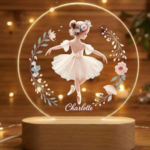 May include: A decorative night light featuring a ballerina illustration. The design includes a ballerina in a white tutu, surrounded by floral accents, and the name "Charlotte". The light sits on a wooden base.