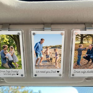May include: Three clear acrylic photo frames clipped to a car sun visor. Each frame displays a family photo with text: "Drive Safe, Daddy! We love you," "We need you, Dad," and "Drive safe, daddy."
