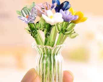 May include: A miniature glass bouquet of colorful flowers in a clear, ribbed vase. The flowers are various colors, including blue, yellow, purple, white, and pink. The stems and leaves are green.