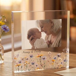 Puede incluir: Marco de fotos de acrílico transparente con una foto de una abuela y un bebé. El marco está decorado con un diseño floral y las palabras "Nanny & Emily". Un pequeño jarrón de flores está a la izquierda.