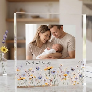 以下が含まれることがあります： 家族写真が入った透明アクリルフォトフレーム。フレームの下部には花の模様があり、「The Smith Family」の文字が入っています。フレームの左側には、小さな花瓶に花が飾られています。