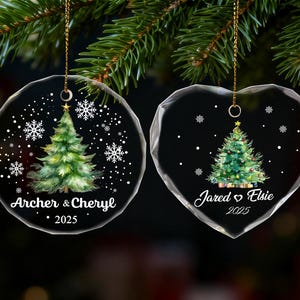 Pu&ograve; includere: Due decorazioni natalizie trasparenti, una rotonda e una a forma di cuore, ciascuna con un disegno ad acquerello di un albero di Natale. L'ornamento rotondo recita "Archer & Cheryl 2025". L'ornamento a cuore recita "Jared & Elsie 2025".