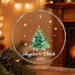 Pu&ograve; includere: Ornamento natalizio rotondo e trasparente con un design ad acquerello di un albero di Natale, fiocchi di neve e i nomi "Hayden ♡ Dinah" e l'anno 2025. L'ornamento &egrave; appeso a una catena d'oro.