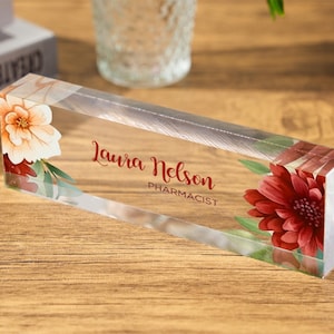 May include: A clear acrylic nameplate with floral accents. The nameplate features the name "Laura Nelson" and the title "Pharmacist" in red script. Floral designs in shades of red, white, and green adorn the edges.