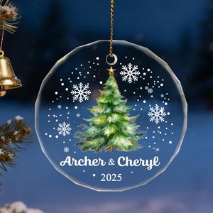 Pu&ograve; includere: Un ornamento natalizio rotondo e trasparente con un design ad acquerello di un albero di Natale, fiocchi di neve e i nomi "Archer & Cheryl" con l'anno "2025". L'ornamento &egrave; appeso a una catena d'oro.