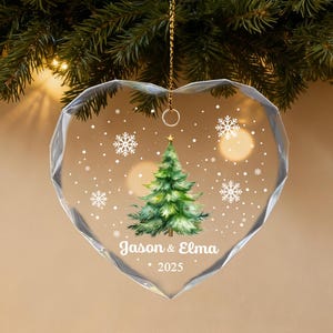 Pu&ograve; includere: Ornamento natalizio trasparente a forma di cuore con un albero di Natale verde dipinto e fiocchi di neve. L'ornamento riporta i nomi "Jason & Elma" e l'anno "2025". L'ornamento &egrave; appeso a una catena d'oro.