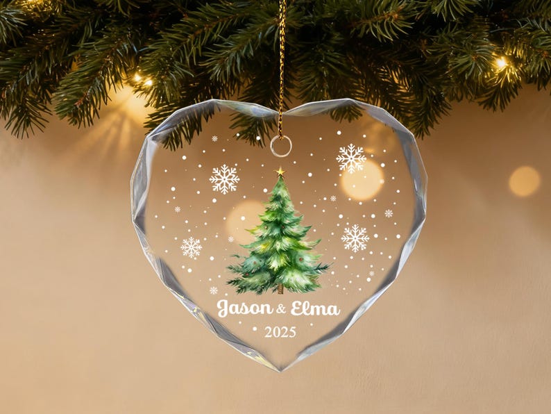 Pu&ograve; includere: Ornamento natalizio trasparente a forma di cuore con un albero di Natale verde dipinto e fiocchi di neve. L'ornamento riporta i nomi "Jason & Elma" e l'anno "2025". L'ornamento &egrave; appeso a una catena d'oro.