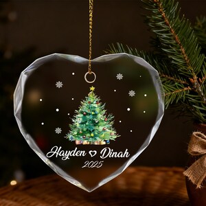 Pu&ograve; includere: Ornamento natalizio a forma di cuore con un design festivo a forma di albero di Natale. L'ornamento in vetro trasparente presenta un albero di Natale verde con ornamenti colorati e i nomi "Hayden & Dinah" e l'anno "2025". Una catena d'oro &egrave; attaccata per appenderlo.