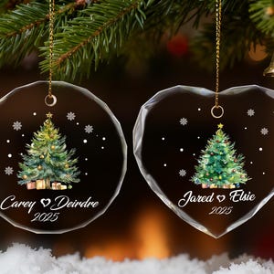 Pu&ograve; includere: Due decorazioni natalizie in vetro trasparente, una rotonda e una a forma di cuore, ciascuna con un disegno di albero di Natale dipinto. L'ornamento rotondo riporta i nomi "Carey & Deirdre 2025" e l'ornamento a forma di cuore riporta i nomi "Jared & Elsie 2025".