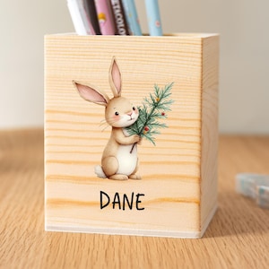 May include: A wooden pencil holder featuring a bunny illustration holding a sprig of greenery. The name "DANE" is printed below the bunny. The holder contains several pens and pencils. The wood has a natural grain.