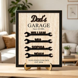 Puede incluir: Un letrero rectangular enmarcado con fondo de veta de madera clara y texto negro que dice Dad's GARAGE Open 24 HRS. Debajo hay cuatro recortes negros con forma de llave inglesa con los nombres WILLIAM, MIA, SOPHIA y NOAH, seguidos de TOYS FIXED FOR FREE EST.2026.