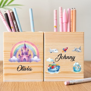 Puede incluir: Dos portalápices de madera con bolígrafos y lápices de colores. Uno presenta un castillo con un arcoíris y el nombre "Olivia". El otro tiene un cohete, un avión, un barco, un velero y el nombre "Johnny".