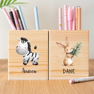 Puede incluir: Dos portalápices de madera con diseños de animales de dibujos animados. Uno presenta una cebra, el otro un conejito sosteniendo una ramita. El nombre "Andrew" está debajo de la cebra y "Dane" debajo del conejo. Los soportes contienen varios lápices y bolígrafos de colores.