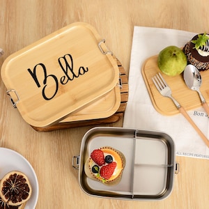 Op de afbeelding: Een bamboe lunchbox met de naam "Bella" in zwart schrift. De doos is open en onthult een roestvrijstalen interieur met verdelers. Een klein dessert met aardbeien en bosbessen zit erin. Een houten snijplank met een cake en fruit is ook aanwezig.