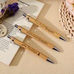 Puede incluir: Tres bolígrafos de bambú personalizados con detalles plateados, cada uno grabado con un nombre: Grayson, Elizabeth y Penelope. Los bolígrafos se muestran sobre un libro, con una pequeña etiqueta de madera que dice "Thank you".