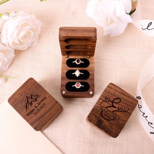 Puede incluir: Una caja de anillos de madera con tres anillos expuestos. La caja está abierta, revelando un interior de terciopelo negro. Hay dos cajas de madera adicionales presentes, una con una montaña y texto, la otra con un monograma y el nombre Martinez.