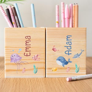 May include: Two wooden pencil holders, one labeled "Emma" and the other "Adam." The "Emma" holder features a purple jellyfish and the "Adam" holder has a blue whale design. Both are filled with colorful pens and pencils.