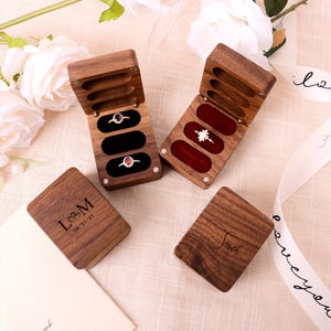 Puede incluir: Cajas de anillos de madera con ranuras de terciopelo abiertas, mostrando anillos con piedras preciosas. Dos cajas están abiertas, revelando los anillos. Dos cajas cerradas tienen iniciales y un nombre grabados. Las cajas están sobre una superficie color crema.