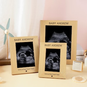 May include: Three light wood picture frames with a textured grain finish. Each frame displays a black and white ultrasound image. The frames are engraved with the text BABY ANDREW at the top and love at first heart beat with a heart icon at the bottom.