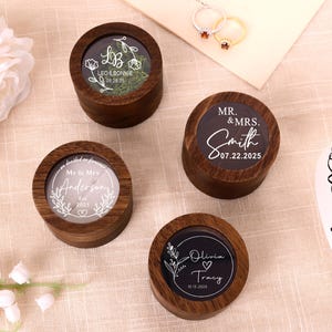 Puede incluir: Cuatro cajas redondas de madera para anillos con tapas transparentes. Cada caja tiene un diseño diferente, incluyendo nombres y fechas. Una caja dice "Leo & Bonnie 09.28.25", otra "Mr. & Mrs. Smith 07.22.2025", una tercera "Mr & Mrs Anderson 2025", y la última "Olivia Tracy 10.15.2025".