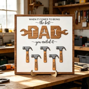 Op de afbeelding: Een vierkant houten bord met een witte achtergrond en de tekst WHEN IT COMES TO BEING the best DAD you nailed it. Het woord DAD is gevormd door houten gereedschapsvormen. Daaronder staan vijf hamerillustraties met de namen Kelly, Daisy, Sarah, Luna en Oliver.