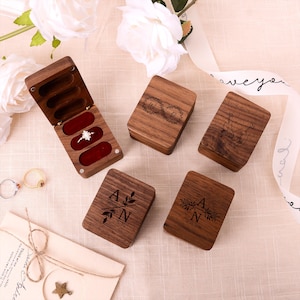 Puede incluir: Cajas de anillos de madera, algunas abiertas con interiores de terciopelo rojo. Las cajas están grabadas con iniciales, diseños florales y fechas. Una caja contiene un anillo de diamantes. Las cajas están sobre una superficie clara.