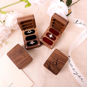 Puede incluir: Cajas de anillos de madera con inserciones de terciopelo, que contienen anillos con piedras preciosas. Dos cajas están abiertas, mostrando anillos. Dos cajas cerradas están grabadas con diseños florales y la letra "A" y el nombre "Stella".