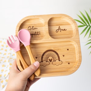 Puede incluir: Plato de bambú dividido en tres secciones, grabado con un diseño de arcoíris, el texto "Guten Appetit!" y el nombre "Ava". Incluye una cuchara y un tenedor de silicona rosa con mangos de madera. Ideal para comidas infantiles.