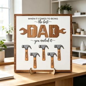 Op de afbeelding: Een vierkant houten bord met een witte achtergrond en de tekst "WHEN IT COMES TO BEING the best DAD you nailed it". Het woord DAD is gevormd door houten gereedschapsvormen. Daaronder staan vijf hamerillustraties met de namen Kelly, Daisy, Sarah, Luna en Oliver.