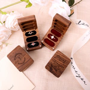 Puede incluir: Cajas de anillos de madera con inserciones de terciopelo, que contienen anillos con piedras preciosas. Las cajas están grabadas con nombres y fechas. Las cajas están abiertas y cerradas, mostrando los anillos. Flores blancas y una cinta en el fondo.