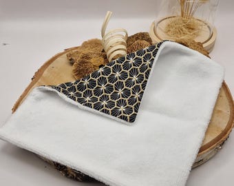 Op de afbeelding: Een witte, zachte washand met een geometrische zwart-gouden hoek. De washand wordt gepresenteerd op een houten schijf met decoratieve elementen op de achtergrond. De algehele esthetiek is strak en modern.