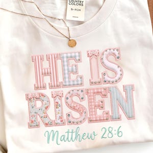 Puede incluir: Camiseta color crema con las palabras "HE IS RISEN" en letras rosas, azules y con estampado floral. El texto "Matthew 28:6" está escrito en una fuente azul claro. También se ven un collar dorado y un sombrero marrón.