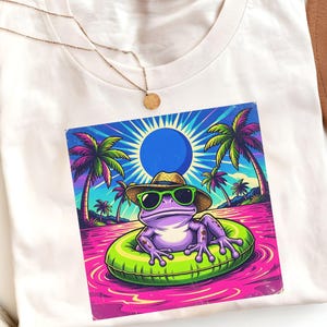 May include: White t-shirt featuring a colorful graphic of a frog wearing sunglasses and a straw hat, floating on an inflatable ring in a pink body of water. Palm trees and a blue sun are in the background. A gold necklace is visible.