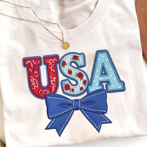 Könnte beinhalten: Weißes T-Shirt mit dem gestickten Wort "USA" in Rot, Blau und Weiß. Das "U" hat ein Paisley-Muster, das "S" Rosen und das "A" Tupfen. Eine blaue Schleife ist darunter gestickt.