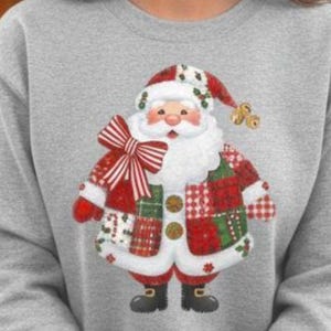 Könnte beinhalten: Ein graues Sweatshirt mit einem Weihnachtsmann-Design. Die Weihnachtsmann-Grafik hat einen weißen Bart, einen roten Anzug mit Patchwork-Design und eine große rote Schleife. Die Person hält einen weißen Becher mit einem rot-grünen Muster.