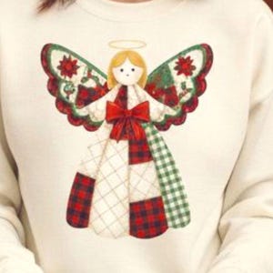 Könnte beinhalten: Cremefarbenes Sweatshirt mit einem Weihnachtsengel-Design. Der Engel hat einen goldenen Heiligenschein, blondes Haar und eine rote Schleife. Die Flügel sind mit roten Weihnachtssternen und grünen Blättern verziert. Das Kleid des Engels ist ein Patchwork aus rotem Karo, Weiß und Grün.