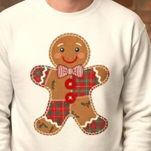 Könnte beinhalten: Ein cremefarbenes Sweatshirt mit einem Lebkuchenmann-Design. Der Lebkuchenmann hat einen rot-grün karierten Körper, eine rote Fliege und zwei rote Knöpfe. Das Design ist auf der Vorderseite des Sweatshirts zentriert.