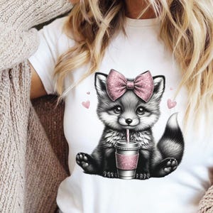 May include: White t-shirt featuring a cute cartoon fox with a pink bow and a pink drink. The fox is black and white with a pink bow and is drinking from a pink cup with a straw. Small pink hearts are scattered around the fox.