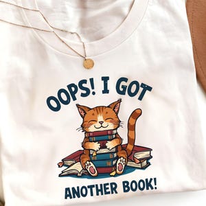 Könnte beinhalten: Weißes T-Shirt mit einer Cartoon-Katze, die auf einem Stapel Bücher sitzt. Die Katze ist orange und weiß und lächelt. Der Text auf dem Shirt lautet: „OOPS! I GOT ANOTHER BOOK!“. Eine goldene Halskette ist sichtbar.