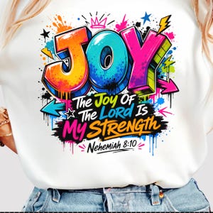 Könnte beinhalten: Ein weißes T-Shirt mit einer bunten Graffiti-Grafik, die das Wort JOY in großen, dreidimensionalen Buchstaben zeigt. Darunter steht der Text The Joy Of The Lord Is My Strength Nehemiah 8:10, umgeben von Farbspritzern, Pfeilen und Sternen.