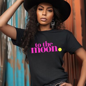 Puede incluir: Camiseta negra con la frase "to the moon" en rosa brillante y un punto amarillo. La modelo lleva un sombrero negro y pendientes plateados. El fondo es una mezcla de azul y naranja.