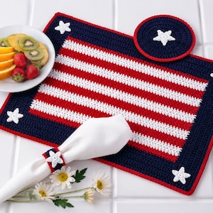 May include: A rectangular crochet placemat and a circular coaster featuring a red, white, and blue striped pattern with white star appliqués. A white napkin is secured with a matching crochet napkin ring featuring a white star appliqué.
