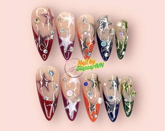Puede incluir: Un conjunto de diez uñas artificiales largas en forma de almendra. Cada uña presenta un diseño de estrella con pedrería plateada y transparente, y un degradado de colores que incluyen rojo, naranja y verde. El texto "Nail by GlassartVN" es visible.